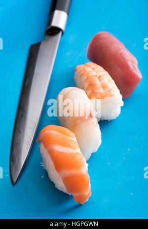 Four different nighiri sushi with slices of raw, fresh, salmon, sea bass, crab and tuna on mounds of rice with a chefs knife alongside, high angle vie Stock Photo