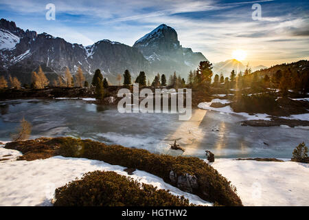 Winter mountain landscape with frozen lake in the front and dramatic sky of evening sun in the background, sunrays protruding trees. Wilderness, raw n Stock Photo