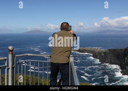 Man taking photograph on mobile phone on edge of cliff at The Fogher Cliffs, Valentia Island, County Kerry, Ireland. Stock Photo
