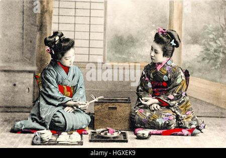 Japan, Vintage postcard hand coloured. Two Geisha in kimono sitting in room conducting tea-ceremony with sweets. Circa 1910. Stock Photo