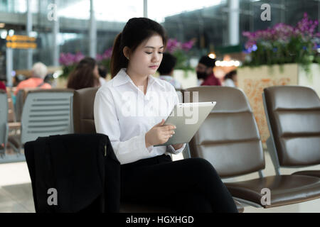 Asian young woman passenger at airport, using her tablet computer while waiting for her flight in terminal at airport. Stock Photo