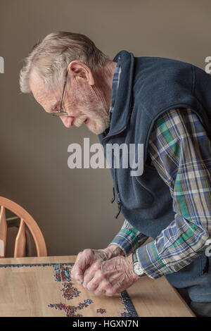 An elderly, bearded man (age 93) wearing eyeglasses and a hearing aid works on a jigsaw puzzle in natural afternoon light. Stock Photo