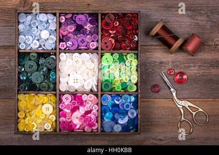A box of colourful buttons, sorted into colour groups, in an old wood box. Spools of thread, a needle and embroidery scissors are placed to one side. Stock Photo