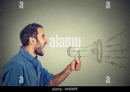 Portrait angry young man holding screaming in megaphone isolated on grey wall background. Negative face expression emotion feeling. Propaganda, breaki Stock Photo