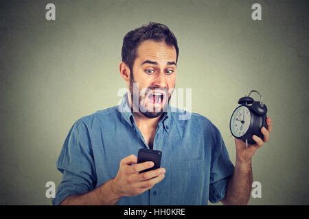 Portrait surprised business man with alarm clock looking at smart phone with funny face expression late for meeting isolated on gray wall background. Stock Photo