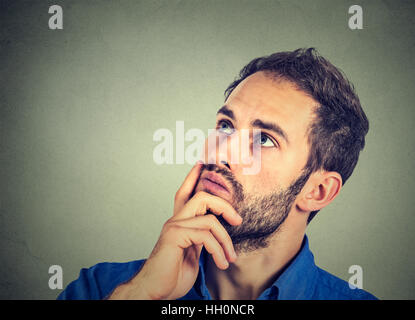 Closeup portrait of a man resting chin on hand thinking daydreaming, staring thoughtfully upwards, copy space to left, isolated on gray wall backgroun Stock Photo