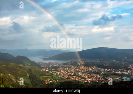 Aerial view of rainbow and cloudy sky over town surrounded with green hills and water Stock Photo