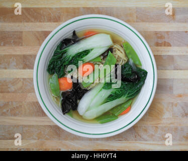 Vegetable noodle soup with bok choy, carrots, snow peas, and wood ear mushrooms in a white bowl on a wooden table. Stock Photo