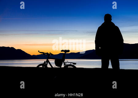 Silhouette of a mountain bike and of a cyclist who is watching the sunset over the lake. Focus on mountain bike Stock Photo