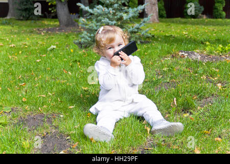 Cute baby girl with mobile phone sitting on green grass Stock Photo
