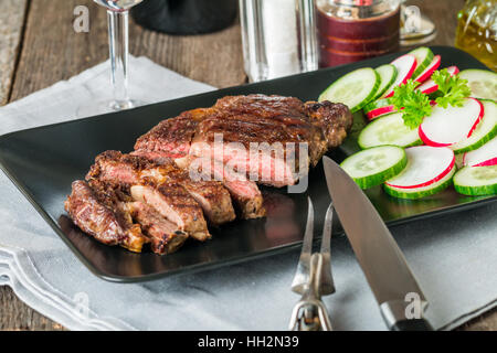 Close up sliced medium rare grilled Beef steak Ribeye with cucumber, radish and red wine on black plate on wooden background Stock Photo