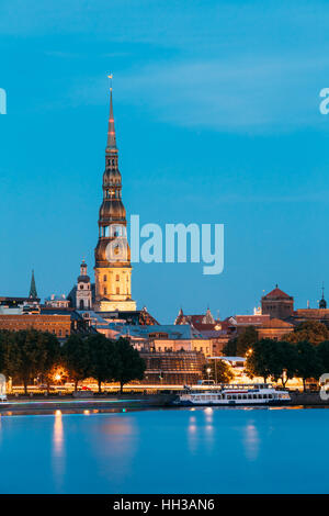 Latvia, Riga Evening Cityscape. The View Of Illuminated St. Peter's Church Tower Behind Daugava River Quay, Old Town In Summer Dusk Under Blue Sky. Stock Photo