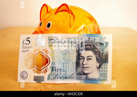 Front of the new polymer UK five pound note leaning against an orange piggy bank. Stock Photo