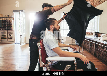 Barber put a sheet to cover his client from cut hair. Male hairdresser at work with handsome man sitting on chair. Stock Photo