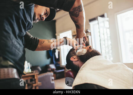 Professional barber shaving customer in his salon. Man getting his beard shaved in a barber shop. Stock Photo