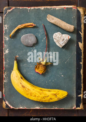 Funny smiley face made with banana, rose and stones on aged grungy book cover Stock Photo