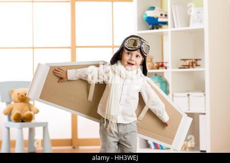 Child boy dressed as pilot or aviator plays with handmade paper wings in his room Stock Photo