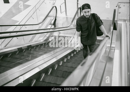young man with a beard in a plaid shirt and a tie and cap, riding on an escalator in the mall Stock Photo