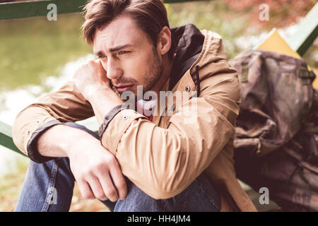 Close-up portrait of young bearded thoughtful man traveler with backpack sitting on pier near river and looking away Stock Photo