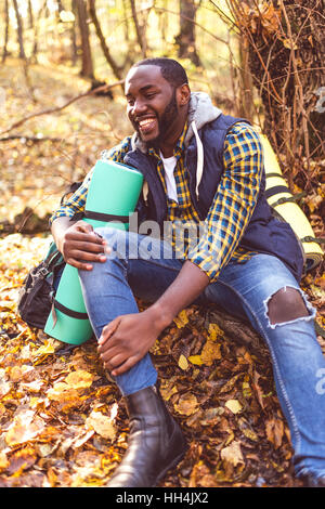 Portrait of handsome smiling man with backpack sitting on tree root and looking away in autumn forest Stock Photo