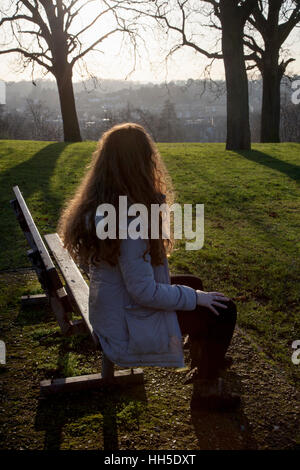 Rear view of a woman sitting alone on a bench in a quiet location with a light sky and trees. Stock Photo