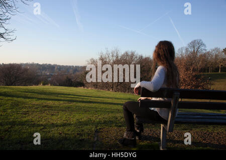 Pensive young woman sitting alone on a bench looking into the distance, in a quiet location with a light sky and trees. Stock Photo