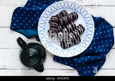 Fragrant cookies of choux pastry in chocolate covered with white decoration on a white plate and teapot on a wooden background. Stock Photo