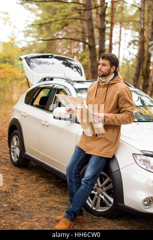 Young bearded man holding map and leaning on white car in autumn forest Stock Photo