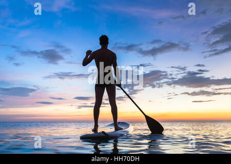 Silhouette of stand up paddle boarder paddling at sunset on a flat warm quiet sea Stock Photo