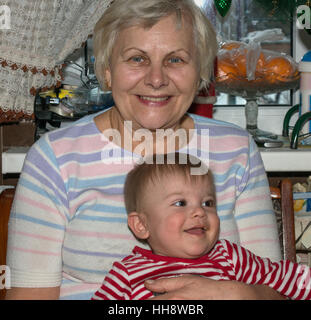 Aged lady cuddles baby boy in red-white striped suit that sitting on her knees just after feeding. Stock Photo