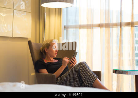 Attractive woman holding and using smart phone while sitting on armchair in modern hotel room. Stock Photo