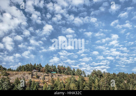 A bright blue sky with many small clouds, above a forested rocky ridge with two homes on its edge. Stock Photo