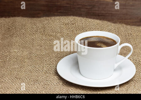 White cup of coffee is standing on the sackcloth. Wooden desk on the background. Stock Photo