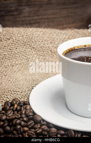 White cup of coffee standing on the sackcloth. Around it are scattered coffee beans and wooden desk is in the background. Vertically. Stock Photo
