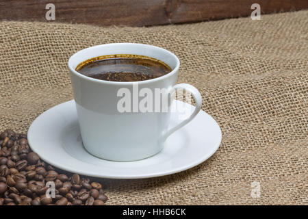 White cup of coffee is standing on the sackcloth. Around it are scattered coffee beans and ground coffee glass. Wooden desk on the background. Stock Photo