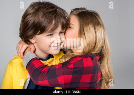 Portrait of schoolgirl hugging and kissing in cheek smiling schoolboy on grey Stock Photo