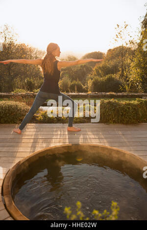 Woman practicing yoga warrior 2 pose on autumn patio with hot tub Stock Photo