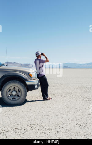 Man standing on vast desert, looking through binoculars and leaning against truck, Black Rock Desert Stock Photo