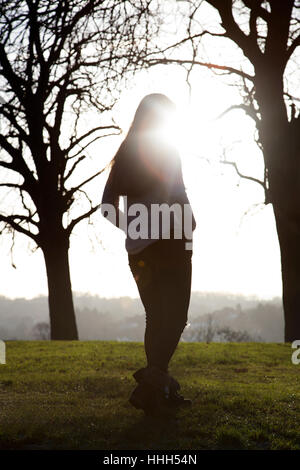 Silhouette of a back view of a unrecognisable woman standing alone by a bench in a quiet location with a sunlight sky and trees. Stock Photo