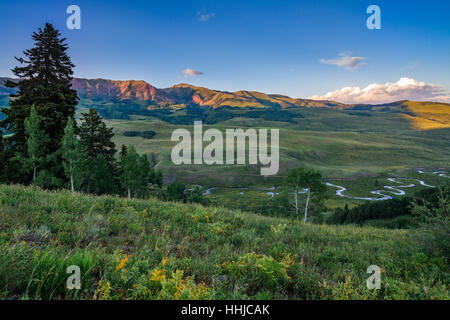 Setting sun makes mountains glow red over green summer fields and a meandering stream near Crested Butte, Colorado Stock Photo
