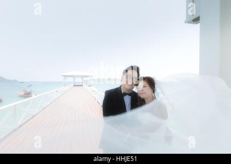 Portrait of cheerful wedding couple hugging and looking at the camera. Asian wedding couple in bridge near sea. Stock Photo