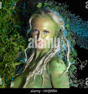 bodypainted woman in front of dark back, partly hidden by green vegetation Stock Photo