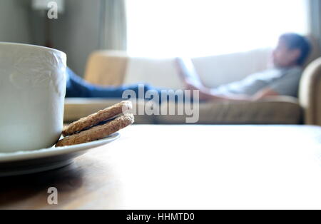 Tea and biscuits or cookies on a wooden coffee table, man relaxing on sofa in blurred background. Space for copy text. Stock Photo