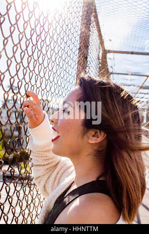 Smiling Asian woman holding chain-link fence on footbridge Stock Photo