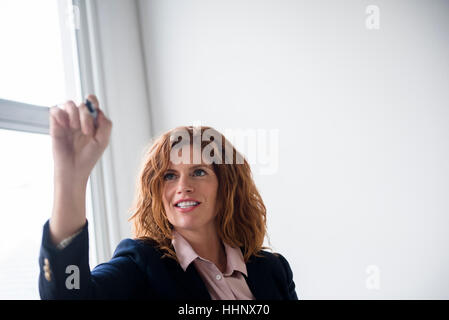 Smiling Caucasian businesswoman writing on glass wall with marker Stock Photo