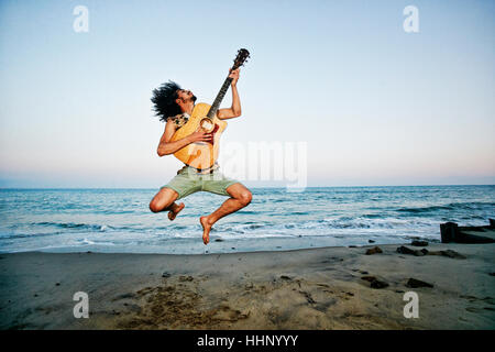 Mixed Race man playing guitar and jumping at beach Stock Photo