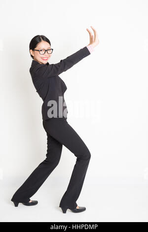 Full body portrait of young Asian businesswoman in formalwear hands pushing on something and smiling, standing on plain background. Stock Photo