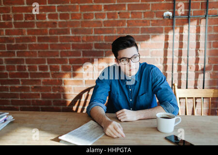 Young man with newspaper, enjoying coffee, sitting at a wooden table in hipster cafe and looking to the side, phone on table Stock Photo