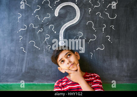 Concepts on blackboard at school. Hispanic boy with doubts and thoughts in class. Portrait of male child thinking against question marks on blackboard Stock Photo