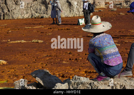 Woman with straw hat and colorful shirt squatting next to rust colored crystal formations in Dallol, Danakil, Ethiopia Stock Photo
